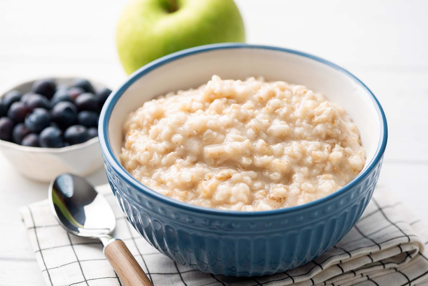 high-protein-carbohydrate-rolled-oats a bowl of cooked rolled oats sitting next to a spoon with blueberries and a green apple in the background